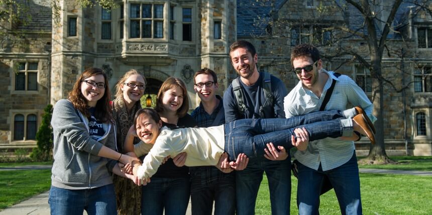 Six smiling students holding a student in horizontally in the air on the quad.
