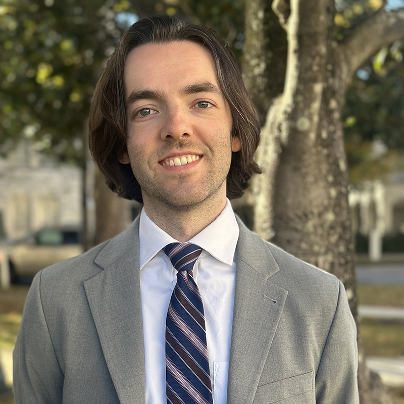 A man in a suit smiles in front of a tree.