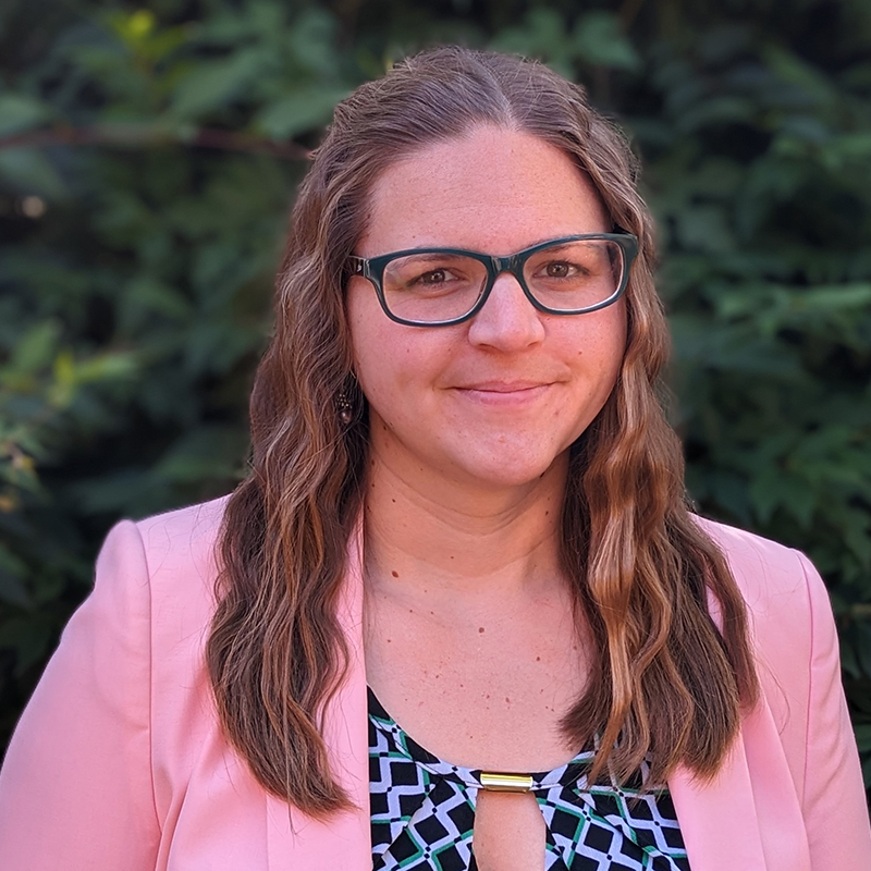 A woman with long brown hair and glasses smiles in front of a shrubbery.