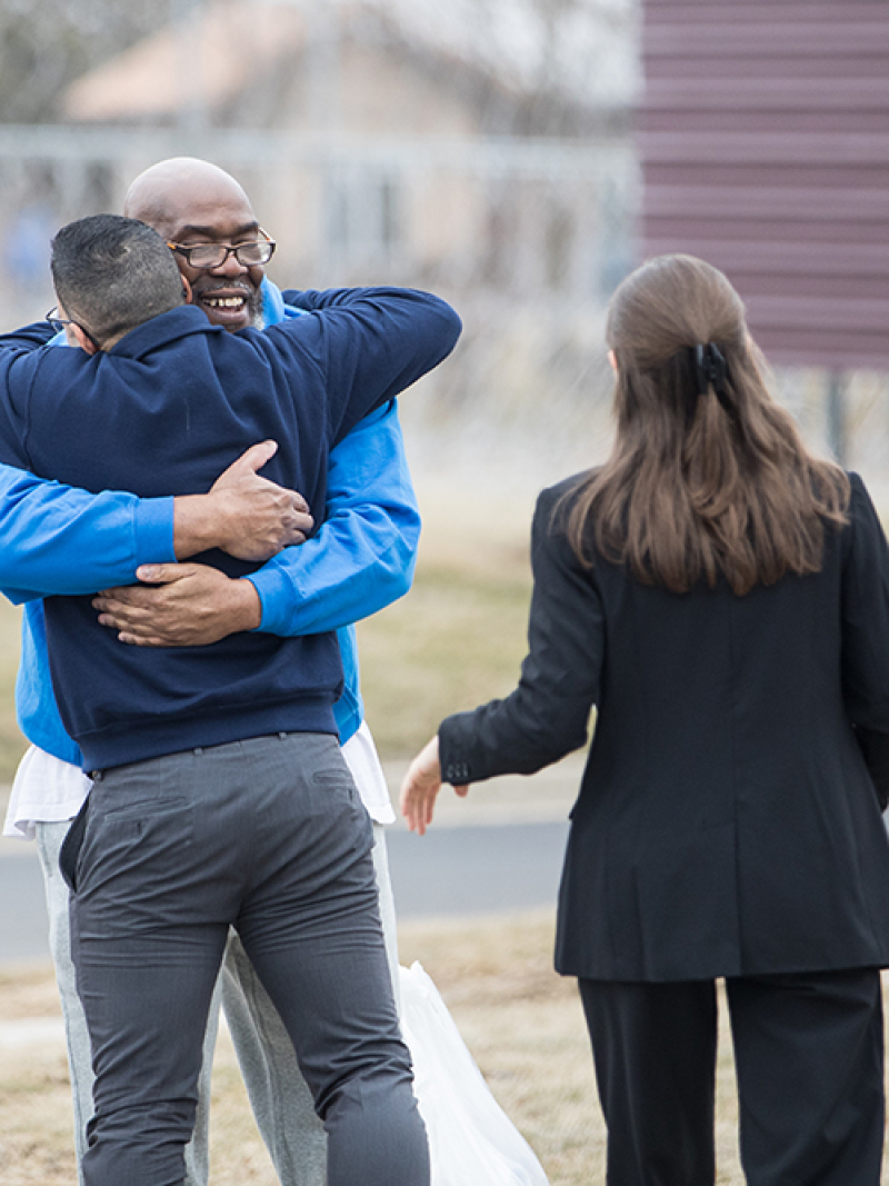 An older man in a hoodie smiles and hugs a younger man on a winter sidewalk.