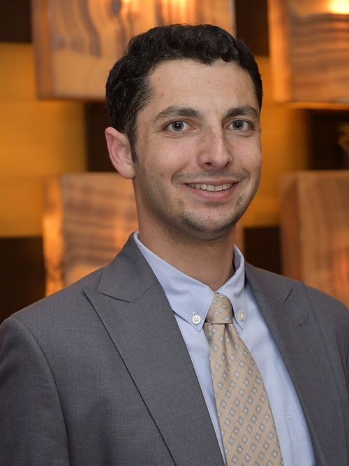 A smiling man in suit and tie stands in front of a colorful restaurant wall.