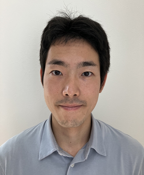 A person with short hair and a light mustache wears a blue button-down shirt.