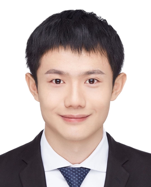 A person with short-cropped hair smiles in a dark suit and tie with a white shirt.