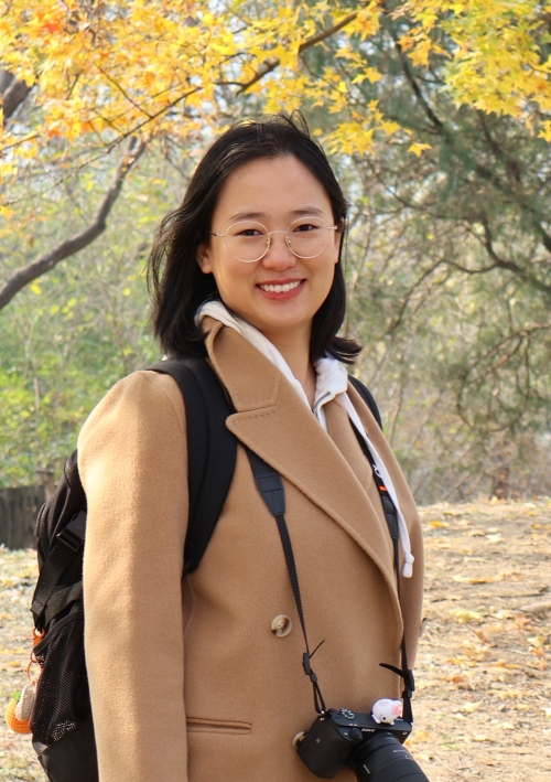 A person in an overcoat and backpack stands in front of a tree in autumn. 