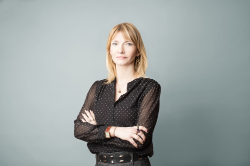A woman with short hair and stylish blouse stands arms folded in front of a blank background.