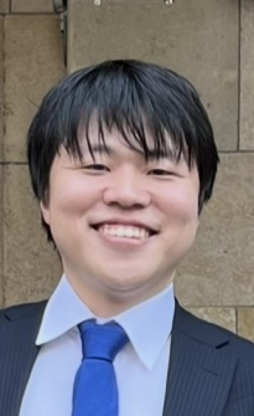 A man in a dark suit smiles in front of a stone wall.