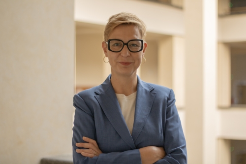 A person with short hair and glasses leans against a column. 