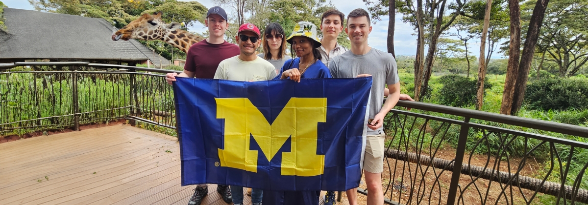 Five individuals stand on a balcony, proudly holding a University of Michigan flag, with a giraffe in the background.