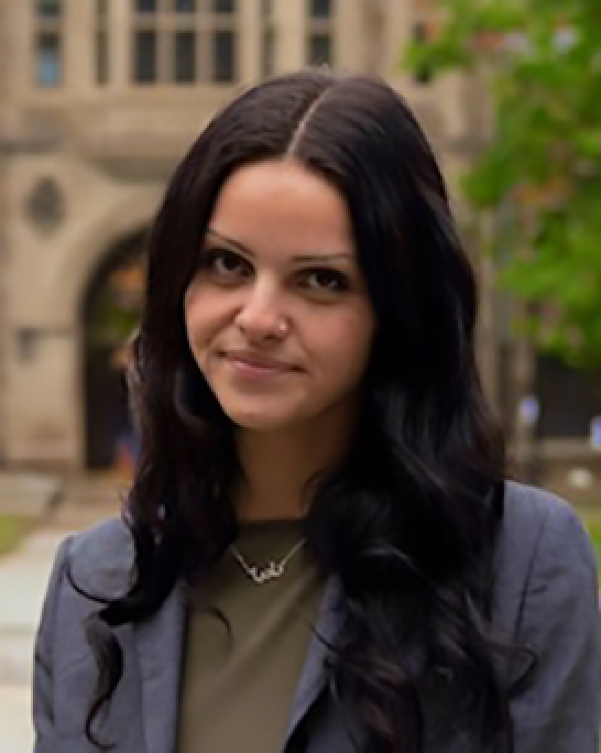 A person with shoulder length hair and a blazer stands in front of a stone building.