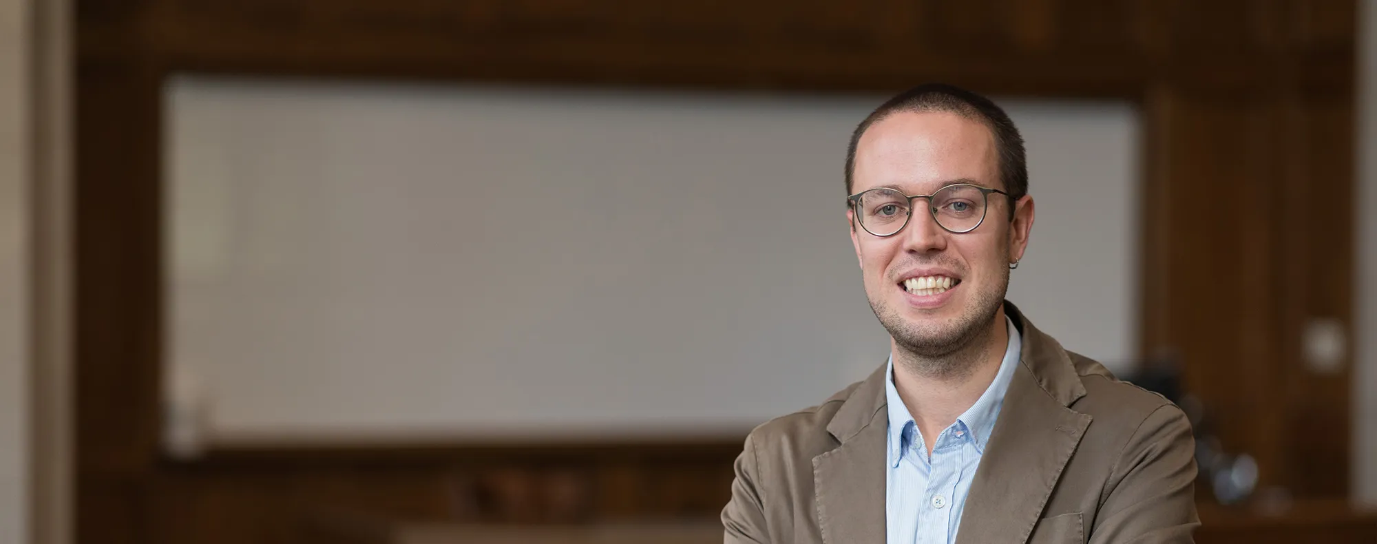 Man, standing at the front of a classroom with a large whiteboard behind him. He is smiling and looking friendly.