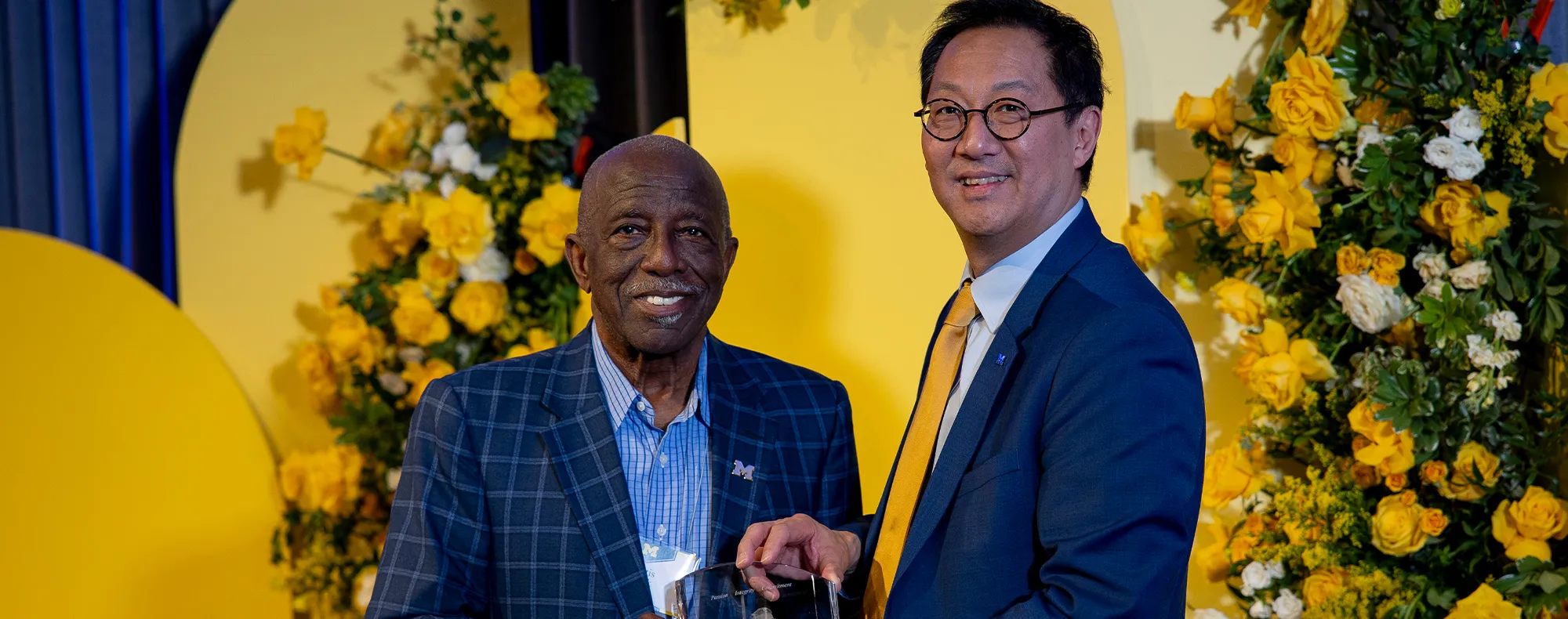 Two people standing in front of a festive background. One person is handing a leadership award to the other person.  