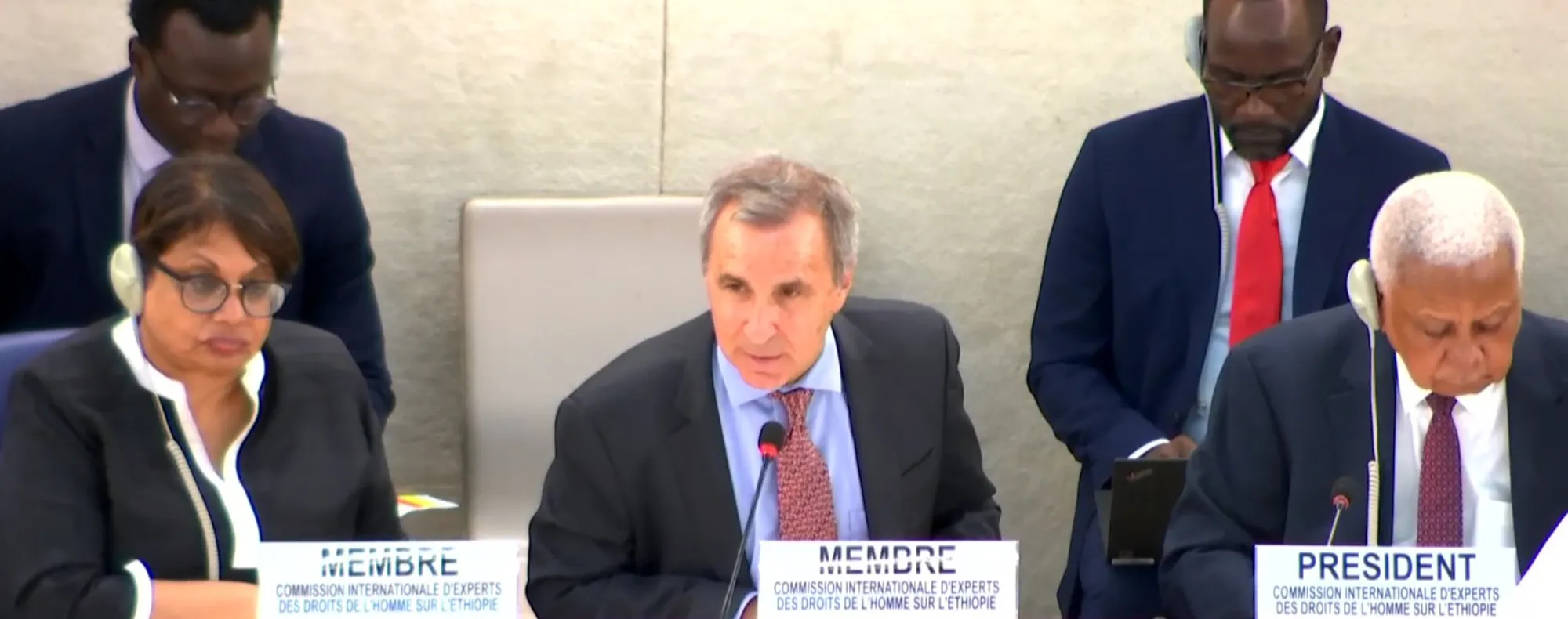 Three people talking on a panel and two people standing in the background