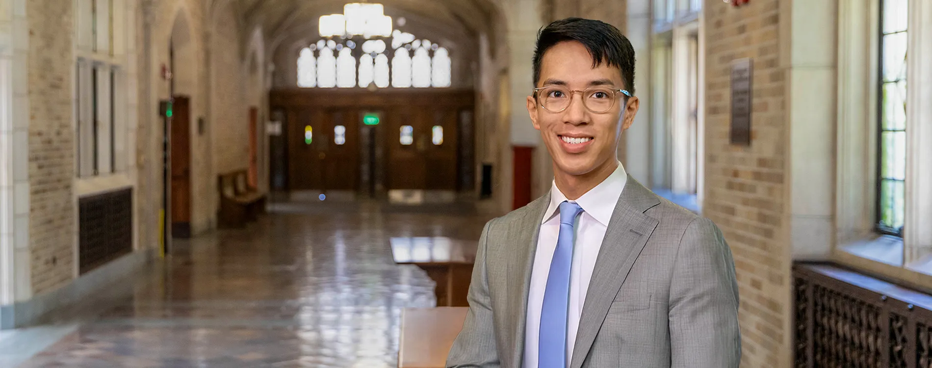 A person wearing glasses, a gray suit, and a light blue tie stands smiling in a long, arched hallway with stone walls and a polished floor, lit by large windows and a hanging light fixture.