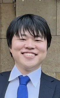 A man in a dark suit smiles in front of a stone wall.