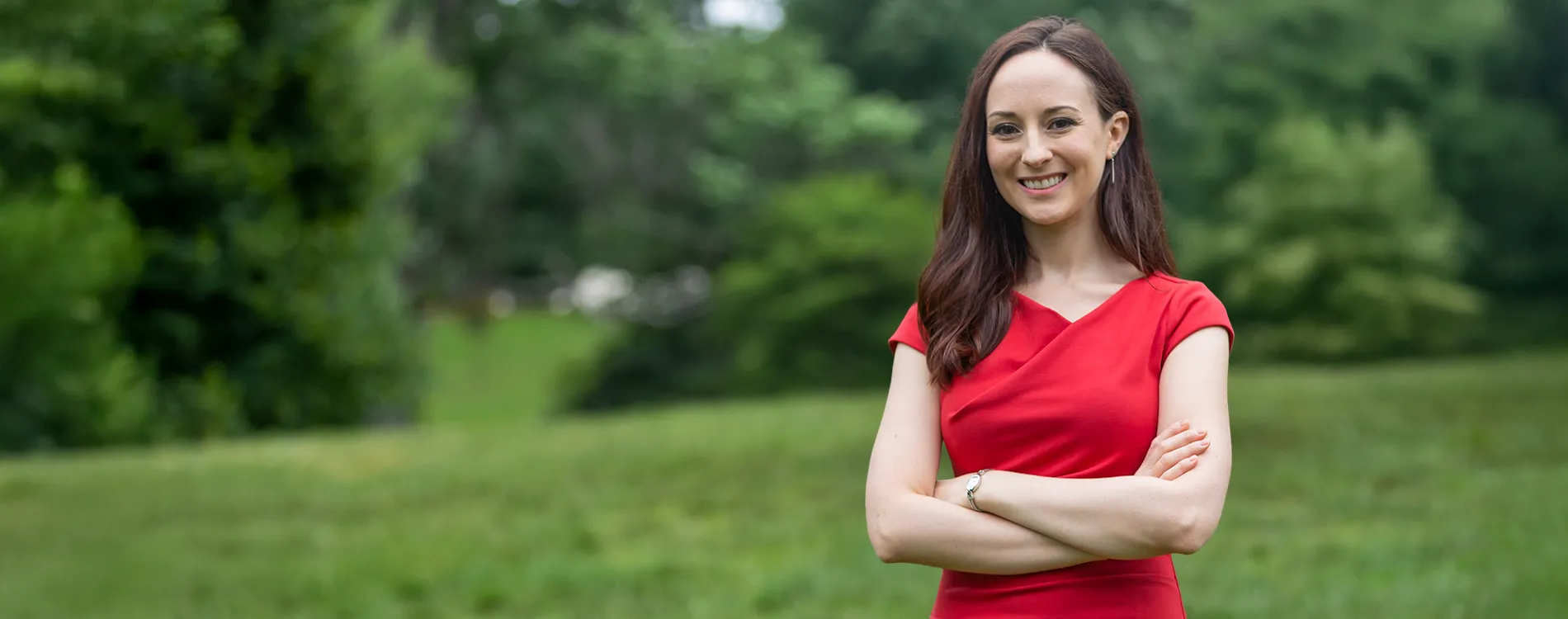 Professor Rachel Rothschild smiling with her arms crossed, standing in a grassy area with trees in the background.