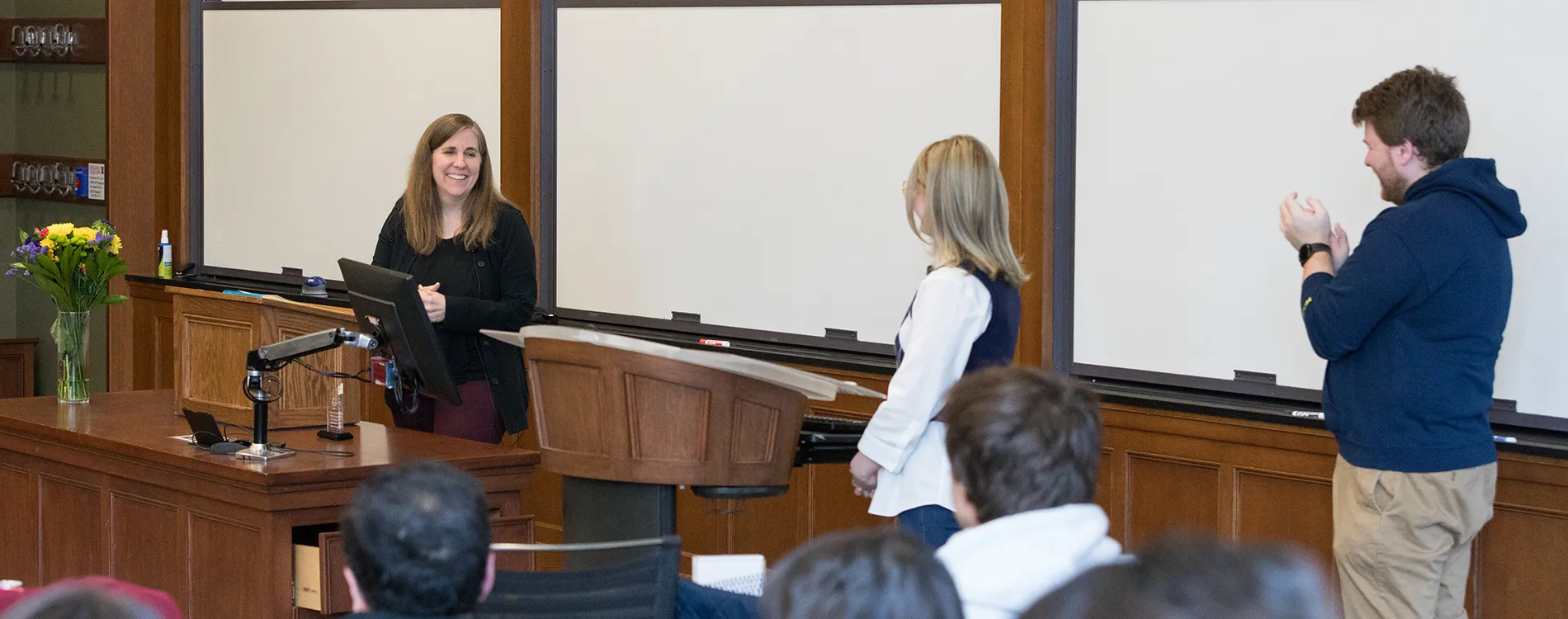 A professor stands behind a lectern while two others stand and clap for her.