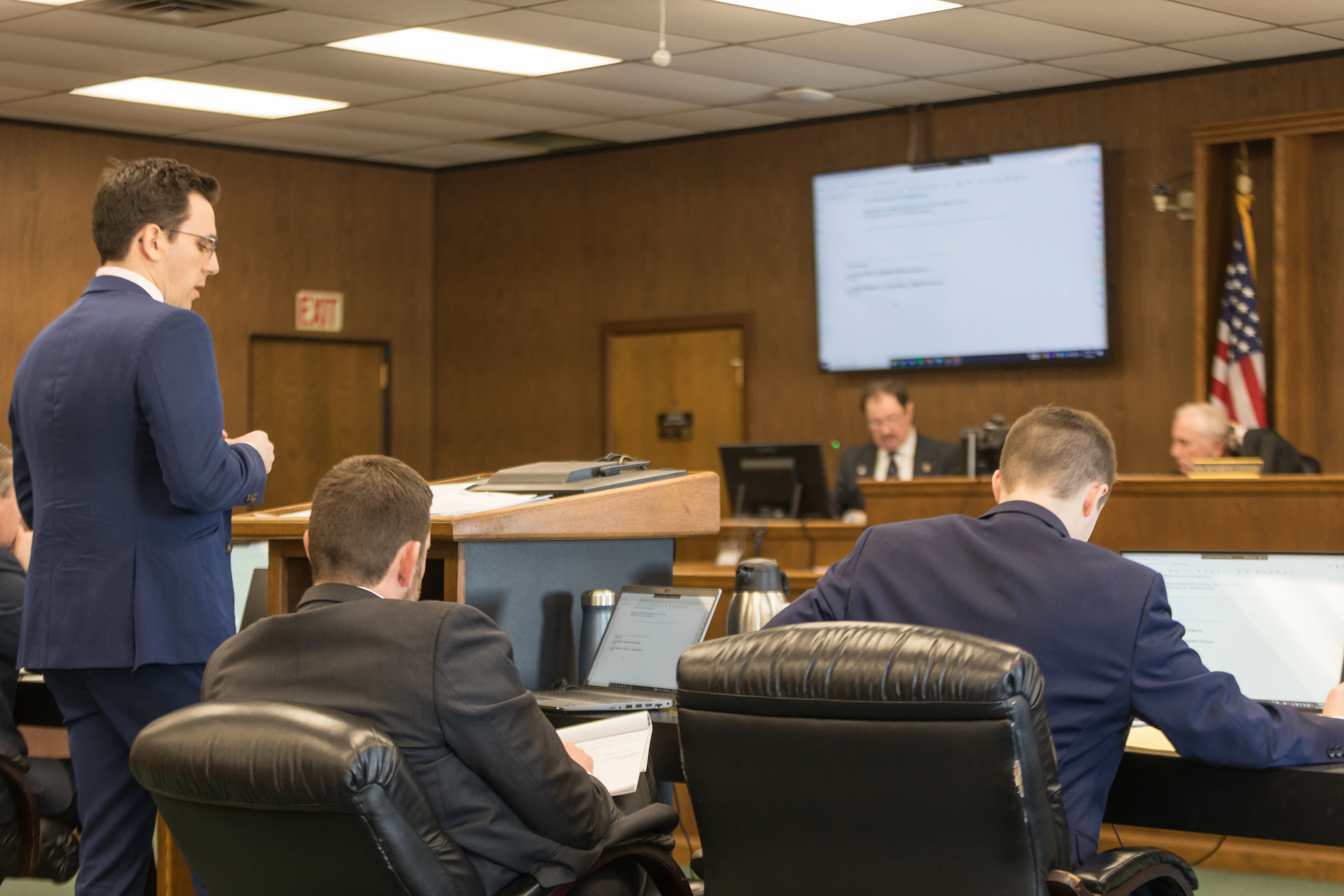 A man in a suit stands in front of a panel of judges in a wood-paneled courtroom.