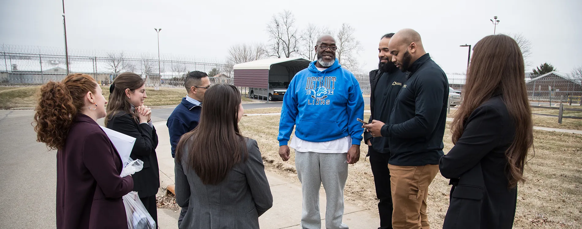 A smiling man in a hoodie stands surrounded by family and friends on a winter sidewalk.