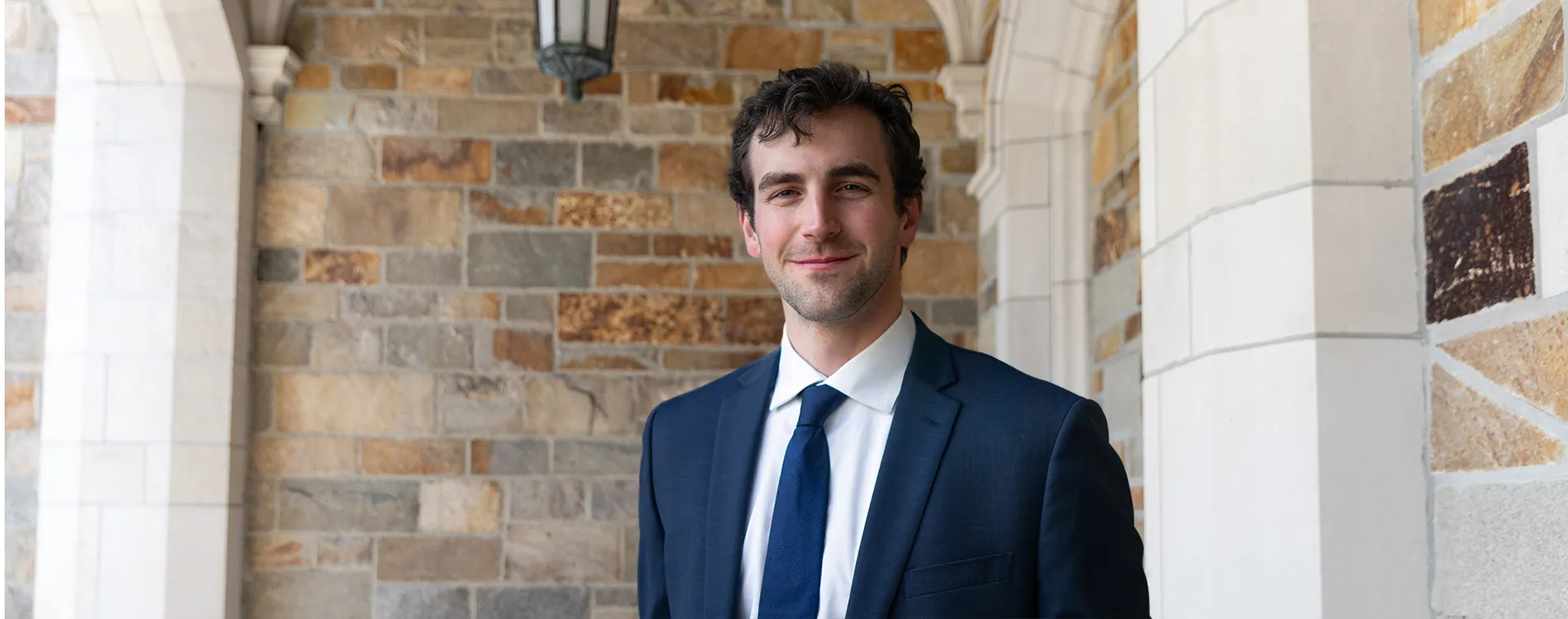 A man in a suit smiles in front of a stone wall.