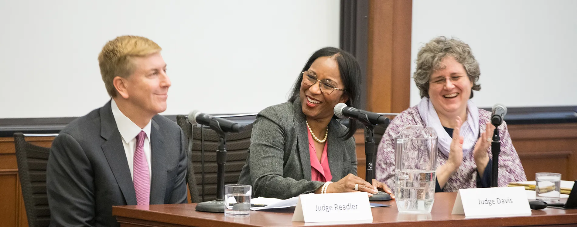 Three people in suits smile at a table with microphones and name placards in front of them. 