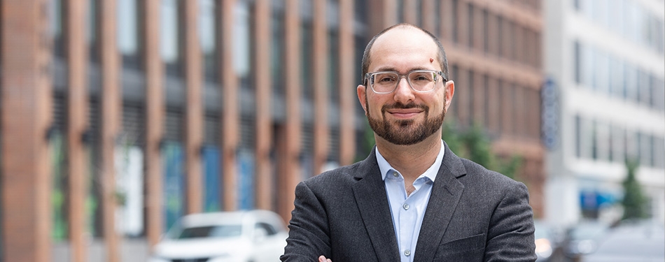 Professor Noah Kazis looking at camera with a blurry city building behind him.