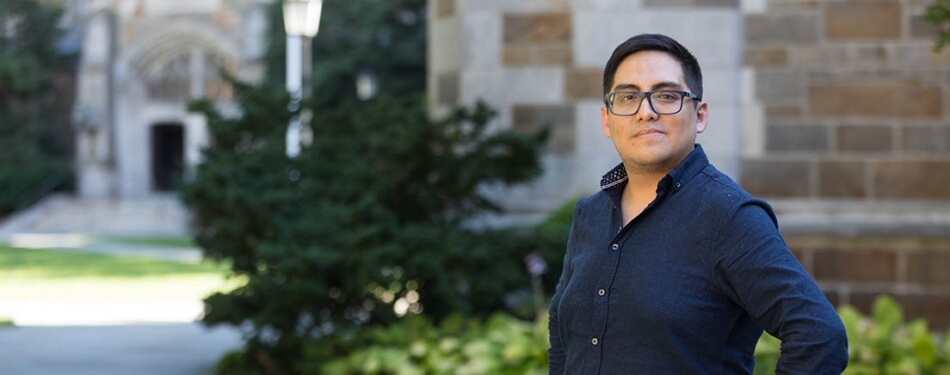 A man in a button down shirt and glasses stands in the Law Quad.