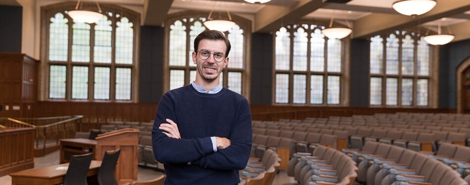 A man in a sweater and glasses stands in a Law School classroom.