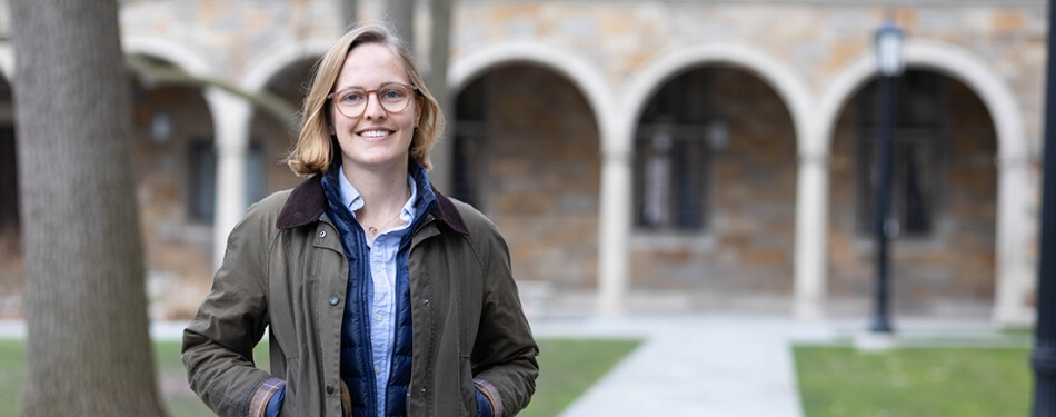 A woman in a raincoat and glasses stands in the Law Quad.