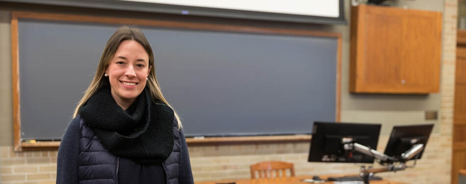 A woman in a dark winter coat and scarf stands in front of a classroom.
