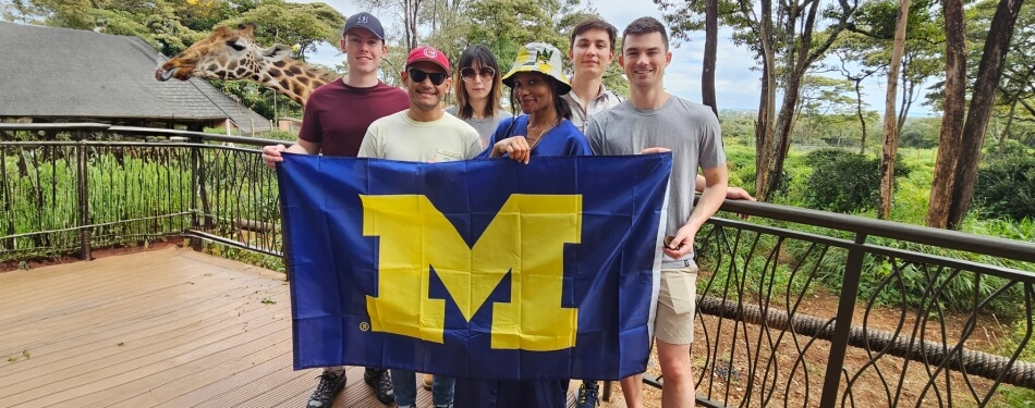 Five individuals stand on a balcony, proudly holding a University of Michigan flag, with a giraffe in the background.