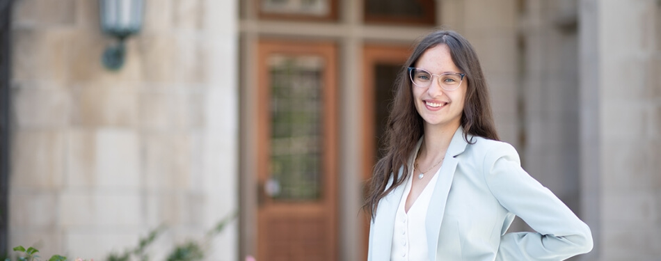 A person with long hair and glasses smiles with her hand on her hip in front of a stone building with wooden doors.