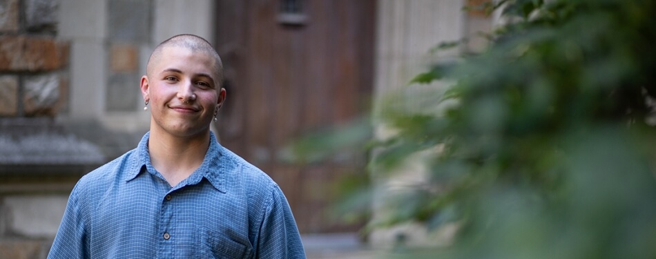 A person with a shaved head and button down shirt stands near a tree in front of a wooden door.
