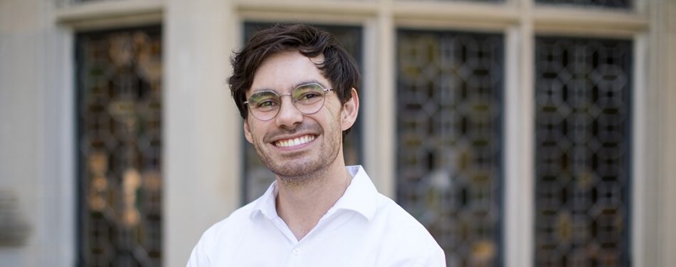 A bespectacled person in a button down shirt smiles in front of a stained glass window.
