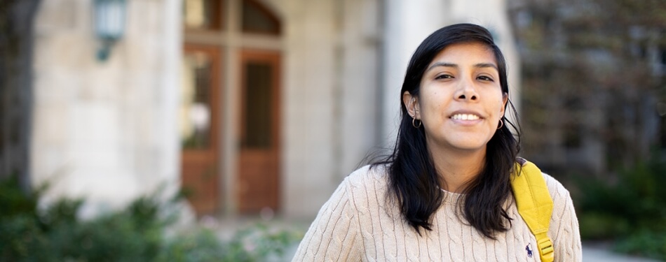 A person with shoulder-length hair and a backpack slung over their shoulder smiles in front of a stone building.