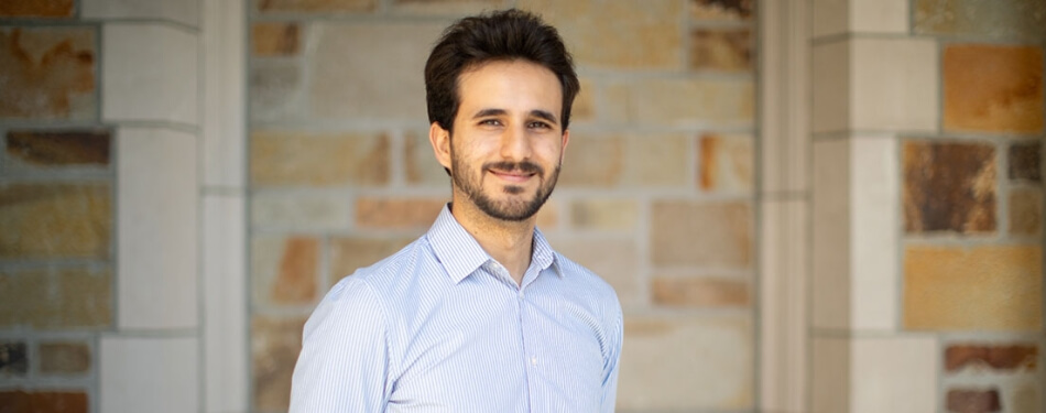 A bearded person smiles in front of a stone wall.