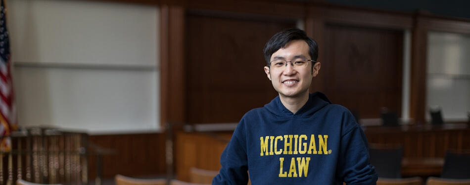 A person in a Michigan Law sweatshirt smiles in front of a classroom marker board..