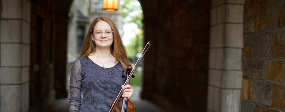 A person with long hair and glasses and holding a violin stands in a stone corridor. 