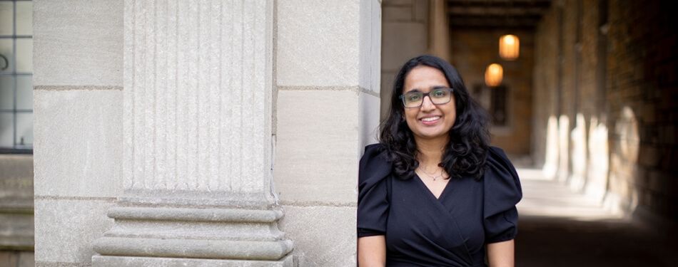 A person with glasses and a blouse leans against a stone pillar in an outdoor passageway.