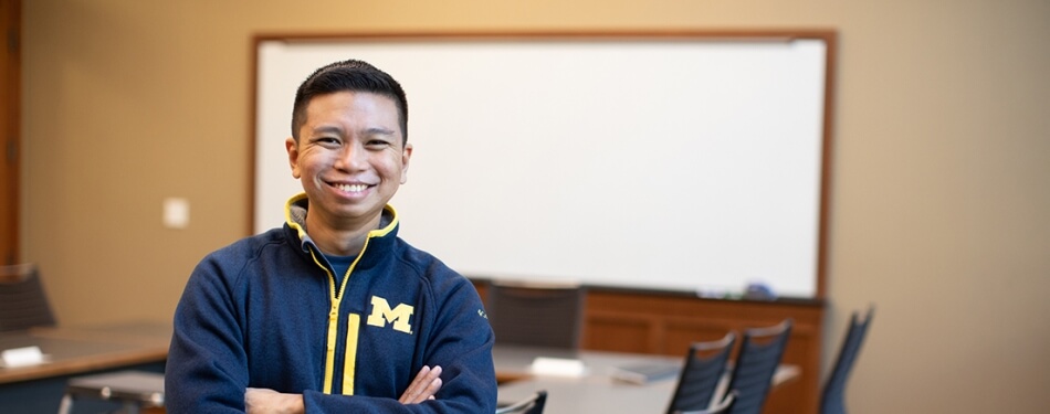 A smiling person with arms crossed stands in front of a classroom markerboard.