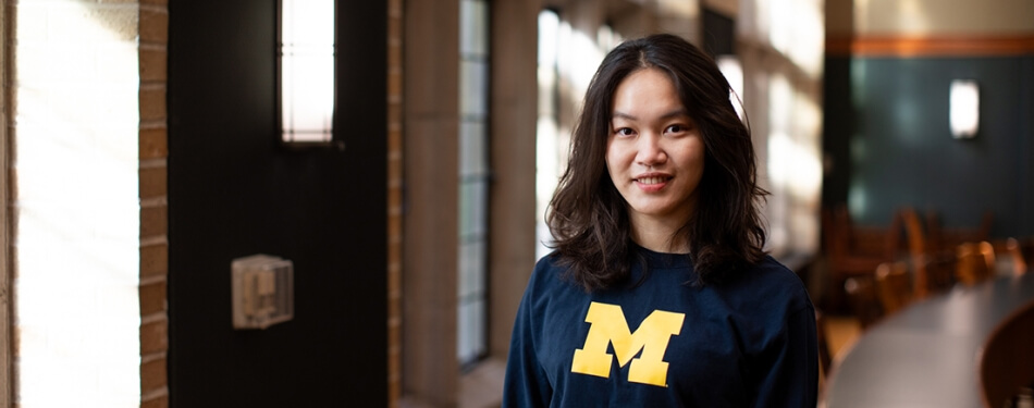 A person in a Michigan shirt with shoulder-length hair smiles in a classroom with sun streaming in the windows. 