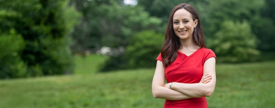 Professor Rachel Rothschild smiling with her arms crossed, standing in a grassy area with trees in the background.