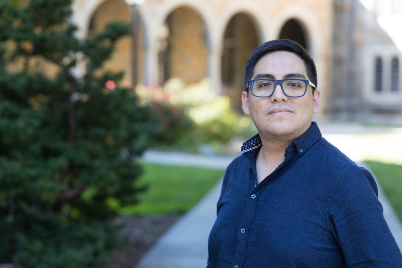 A man in a button down shirt and glasses stands in the Law Quad.