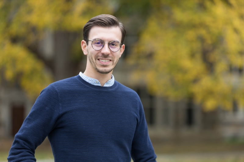 A man in a sweater and glasses stands in front of autumn trees.