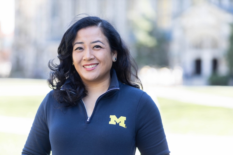 A woman in a Michigan pullover stands in the sunshine on the Law Quad.