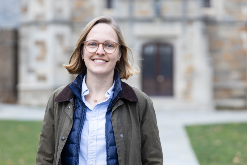 A woman in a raincoat and glasses stands in the Law Quad.