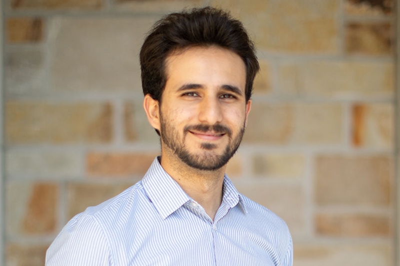 A bearded person smiles in front of a stone wall.