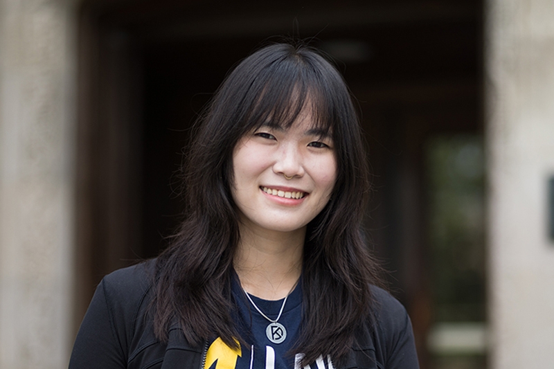 A person with long, dark hair smiles in front of a stone corridor.