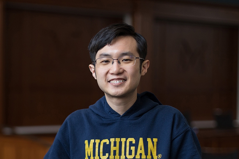 A person with short hair and glasses smiles in front of a wood-paneled wall. 