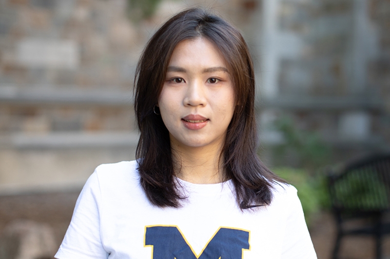 A person with shoulder-length hair stands in front of a stone facade.