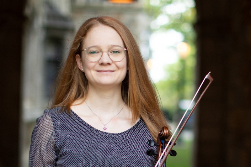 A person with long hair and glasses and holding a violin stands in a stone corridor.